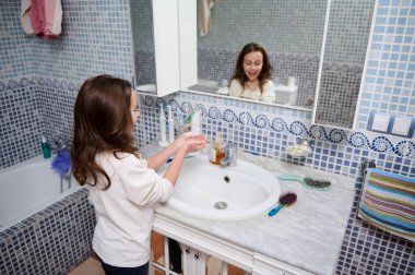 Genç bir kız mermer bir banyo lavabosunun önünde ellerini yıkıyor. Parlak mavi mozaik fayanslar, bir havlu, fırça, tarak ve sabun dağıtıcısı aynaya yansıyor..