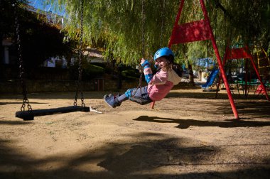 Pembe ceketli ve mavi kasklı genç bir kız güneşli bir günde parkta salıncakta salıncaktan keyif alıyor..