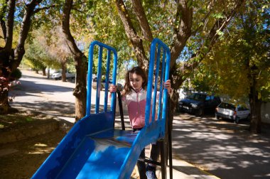 Genç bir kız gölgeli bir parkta, açık havada ağaçların ve arka planda park edilmiş arabaların arasında parlak mavi bir oyun parkına tırmanıyor..