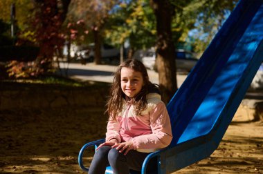 Pembe ceketli, gülümseyen bir kız güneşli bir sonbahar parkında mavi bir oyun parkında oturmuş aile ve açık hava yaşam tarzı için ideal sıcak, neşeli bir çocuk anı yakalıyor..