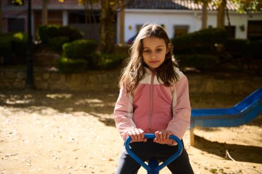 Genç bir kız, pembe ve krem rengi ceket giymiş mavi bir baharda ata biniyor. Ağaçlı güneş parkı ve arka planda bir kaydırak, kaygısız çocukluk anılarını yakalıyor..