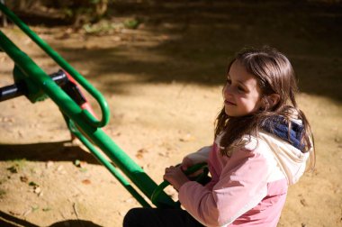 Pembe ceketli genç bir kız güneşli bir parkta tahterevallide oturuyor, gülümsüyor ve neşeli bir anın tadını çıkarıyor..