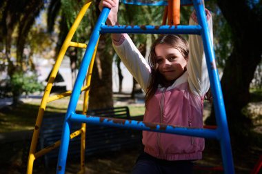 Pembe ceketli bir kız, güneşli bir günde parkta oynarken gülümseyen mavi ve sarı bir tırmanma parkına tırmanıyor..