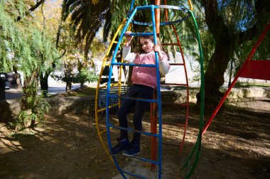 Genç bir kız güneşli bir parkta, parlak metal bir tırmanma parkına tırmanıyor, ağaçların arasında, çok renkli bir yapıda eğleniyor..