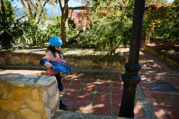 Pembe ceketli ve mavi kasklı genç bir kız, güneşli bir parkta elinde mavi bir kaykayla taş bir duvarın yanında duruyor. Güvenli oyun alanı. Açık hava aktivitesi ve çocukluk eğlencesi..