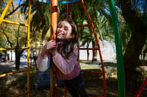 Gülümseyen bir kız güneşli bir parkta, parlak bir tırmanma parkına tutunur, kaygısız açık hava oyunlarını ve gençlik neşesini yakalar..