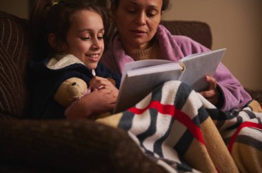 Kanepeye kıvrılıp, sıcak bir battaniyenin altında yanında oyuncak bir ayıyla kitap okuyan anne ve genç bir kızın samimi bir sahnesi..