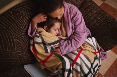 Bir anne ve çocuk yumuşak bir battaniyeye sarılmış bir kanepede sarılır. Evdeki sıcak, samimi an sevgi, ilgi ve aile rahatlığını iletir..