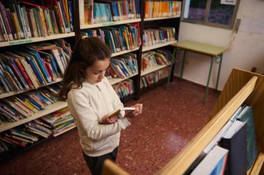 Genç bir kız kütüphanede duruyor, raftan kitap okuyor, sessiz ve çalışkan bir ortamda öğrenme ve keşfetme işine dalmış..