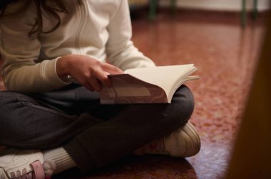 Genç bir öğrenci fayanslı zeminde bağdaş kurup kitap çeviriyor. Sahne, eğitim ortamında öğrenme, odaklanma ve merak taşır.