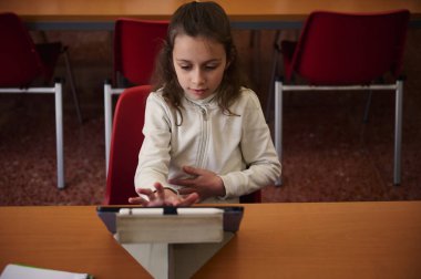 Genç bir kız, okul kütüphanesindeki ahşap bir masada kırmızı sandalyelerle çevrili bir tablete odaklanıyor. Bir sınıfta öğrenme, konsantrasyon ve dijital eğitimi yakalar..
