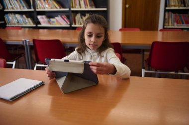 Genç bir kız çalışmak için bir tablet kullanarak kütüphane masasında oturuyor. Kitap rafları ve defterler etrafını sarmış, okula dönüş öğrenimini ve okuma sevincini yakalıyor..