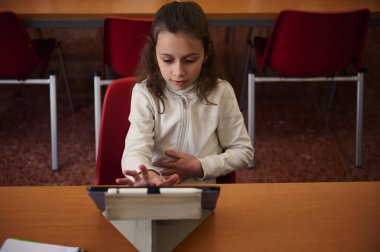 Genç bir kız, okul kütüphanesindeki ahşap bir masada kırmızı sandalyelerle çevrili bir tablete odaklanıyor. Bir sınıfta öğrenme, konsantrasyon ve dijital eğitimi yakalar..