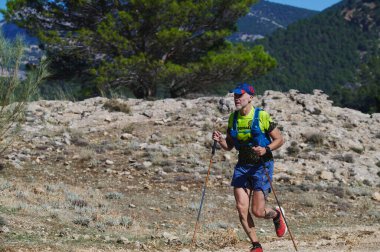 18 Ekim 2025 - Quesada, İspanya - Maraton Sierra Cazorla Patikası Engebeli bir dağ manzarasının ortasında yürüyüş direkleri ile kayalık, güneşli bir patika boyunca neon vites gücü ile koşan bir erkek..