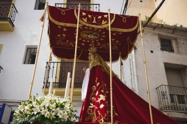 Quesada, Endülüs, İspanya. 18 Nisan 2024. Altın işlemeli kırmızı kadifeye sarılı törensel bir şamandıra, yerel bir festival sırasında beyaz çiçeklerin arasında taçlandırılmış bir heykel taşıyor..