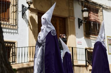 Quesada, Endülüs, İspanya. 18 Nisan 2024 Mor cüppeli üç kukuletalı şahıs, yerel bir tören sırasında güneşli bir caddede yürürken gizemli, törensel bir sahne yaratıyor..