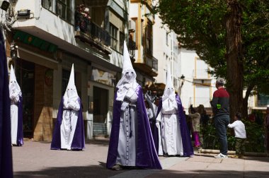 Quesada, Endülüs, İspanya. 18 Nisan 2024. Mor cüppeli tövbe edenler, izleyiciler İspanya 'da geleneksel ve resmi bir festival sahnesini çekerken güneşli bir sokakta yürürler..