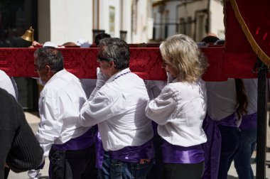 Quesada, Endülüs, İspanya. 18 Nisan 2024. Beyaz gömlekli ve mor kuşaklı bir grup insan, geçit töreni sırasında kırmızı bir dekoratif şamandıra taşıyor. Bu, toplumsal gelenek ve festival ruhunu yansıtıyor..