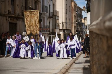 Quesada, Endülüs, İspanya. 18 Nisan 2024. Beyaz ve mor cüppeli çocuklar festival sırasında pankartla yürüyorlar, komşular güneşli bir cadde boyunca izliyorlar.