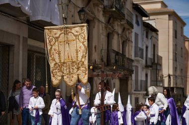 Quesada, Endülüs, İspanya. 18 Nisan 2024. Yerel bir festival sırasında, ruhban sınıfı ve mor cüppeli çocukların bayrak ve ahşap haç taşıdığı dini bir geçit töreni..