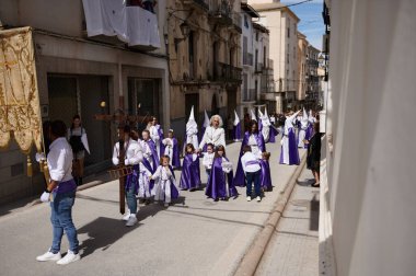 Quesada, Endülüs, İspanya. 18 Nisan 2024. Mor cüppeli çocuklar ve yetişkinlerle dolu açık hava dini geçit töreni, eski binalar, gelenekler ve törenlerle dolu güneşli bir cadde boyunca yürüyorlar..