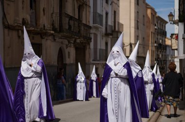 Quesada, Endülüs, İspanya. 18 Nisan 2024. Beyaz cüppeli ve mor pelerinli tövbe edenler kutsal hafta boyunca güneşli bir caddede yürürler..