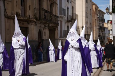 Quesada, Endülüs, İspanya. 18 Nisan 2024. Geleneksel bir tören sırasında beyaz cüppeli ve mor pelerinli adamlar dar bir sokakta yürürler..