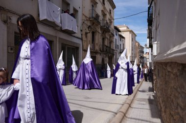 Yerel bir festival sırasında, Endülüs 'ün Quesada kentindeki güneşli bir cadde boyunca mor pelerinli kapüşonlu katılımcıların geçit töreni. Meraklı izleyiciler ve balkonlar dar yolda. Quesada, Endülüs, İspanya. 18 Nisan 2024