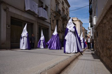 Quesada, Endülüs, İspanya. 18 Nisan 2024. Mor pelerinli beyaz kapüşonlu adamlar dar bir sokakta yürürken kutsal hafta geçidi gözetleyen insanlar, parlak gün ışığı, geleneksel tören atmosferi..