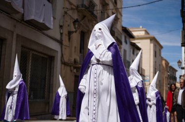 Mor pelerinli beyaz cüppeli kapüşonlu tövbeklerin geçit töreni Quesada, Endülüs, İspanya 'da dar bir cadde boyunca ilerliyor. 18 Nisan 2024