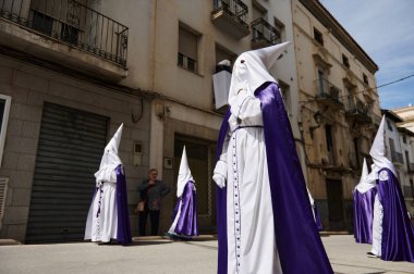 Quesada, Endülüs, İspanya. 18 Nisan 2024. Beyaz cüppeli ve mor pelerinli başlıklı şahıslar, eski binalara karşı kontrastı yakalayan geleneksel bir dini etkinlik olan sokak gösterisine katılıyorlar..