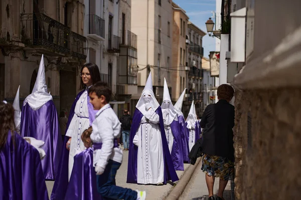Quesada, Endülüs, İspanya. 18 Nisan 2024. Beyaz cüppeli ve mor pelerinli sokak katılımcıları, güneşli tarihi bir kasabada seyircilerin yanından geçen kapüşonlu figürler..