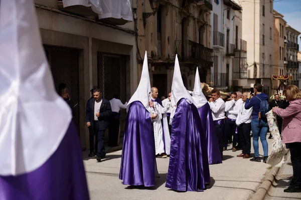 Quesada, Endülüs, İspanya. 18 Nisan 2024. Güneşli bir Akdeniz kasabası boyunca fotoğraf çeken mor ve beyaz giysili katılımcıların yer aldığı bir sokak geçidi..