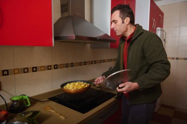 Modern bir mutfak sobasının yanında duran bir adam kızartma tavasında patates kaması pişiriyor. Tavayı, kırmızı dolapları ve arka planda seramikleri çevirirken cam bir kapak tutuyor..