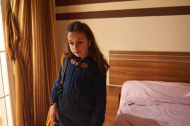 Küçük kız çantasını taşıyor, okula hazırlanıyor, sıcak odasının içinde duruyor.