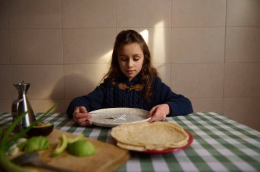 Küçük bir kız yemek masasında otururken kahvaltı yapıyor. Yemek masası yeşil kareli bir bezle kaplıdır. Ekmek ve zeytinyağlı avokado masada. Pencereden gelen güneş ışığı nazikçe yüzüne düşer..