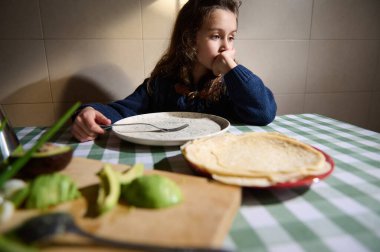 Düşünceli küçük kız yemek masasında oturuyor, rüyasında gözlerini kaçırıyor. Yemek masası yeşil kareli bir bezle kaplıdır. Pencereden gelen güneş ışığı nazikçe yüzüne düşer..