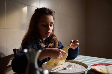 Küçük kız sabah kahvaltı yapıyor, mutfakta yemek masasında oturuyor, elinde ev yapımı ekmek tutuyor.