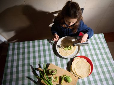 Yukarıdan: güzel bir kız çocuğu avokadolu bir ekmeğin üzerine zeytinyağı döküyor, Akdeniz kahvaltısının tadını çıkarıyor. Mutfak penceresinden yüzüne güneş ışınları düşüyor.