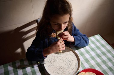 Avokado ve salatalı rulo ekmeği ısıran beyaz tatlı bir kız, sıcak bir evde yemek masasında oturuyor..