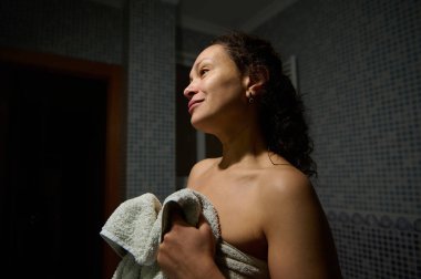 Banyo havlusuna sarılı, duştan sonra banyoda duran, elinde havlu tutan ve aynanın yansımasına bakıp gülümseyen kendine güvenen güzel bir kadının özgün portresi.