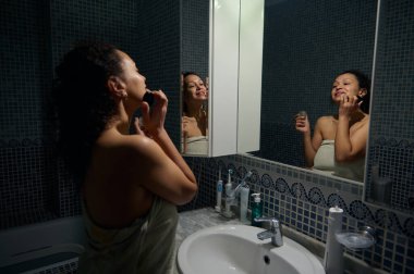 Aynaya yansıyan bir kadın dudaklarına nemlendirici serum sürüyor, banyoda lavabonun yanında duruyor, banyo havlusuyla sarılmış. Kaplıca, güzellik prosedürleri, cilt ve vücut bakımı konsepti