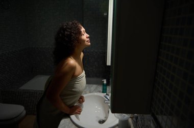 Kıvırcık saçlı, orta yaşlı, banyo havlusuna sarılmış, lavabonun yanında duran, aynadaki yansımasına hayran olan kadın. Doğal güzellikte bir kadın günlük hijyen ve güzellik rutininin tadını çıkarıyor.