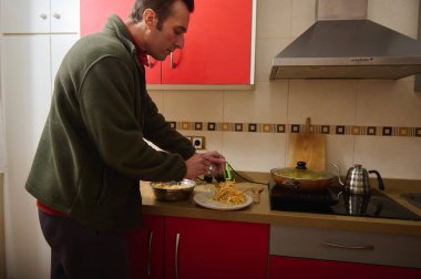 Yeşil ceketli bir adam çağdaş bir mutfakta yemek hazırlar. Fırında bir tencere kaynarken makarnanın üzerine peynir serpiyor..