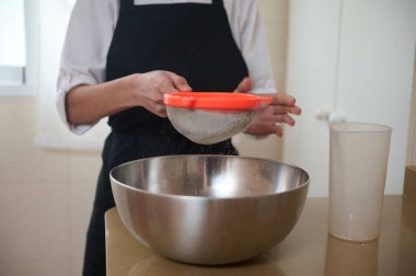 Beyaz gömlekli ve siyah önlüklü bir insan, unu, güneşli bir mutfak tezgahında hamuru hazırlayan paslanmaz bir kâseye yerleştirir..