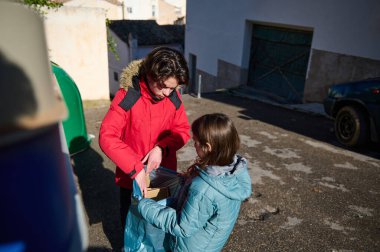 Kırmızı kışlık ceketli bir kadın açık hava avlusunda açık mavi ceketli bir kıza mavi bir çanta veriyor..