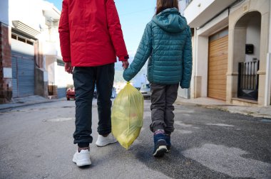İki kişi ellerinde parlak sarı bir çantayla bir şehir caddesinde yürüyor. Aile takım çalışmasının, günlük sorumluluğun ve şehir hayatının altını çizen sıradan bir açık hava anı.