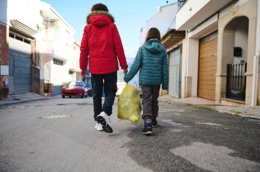 İki çocuk, sarı bir çanta taşıyarak sakin bir şehir caddesinde el ele yürüyorlar. Kışlık ceketler giyiyorlar. Sıradan, günlük bir aile anı gibi görünüyor..