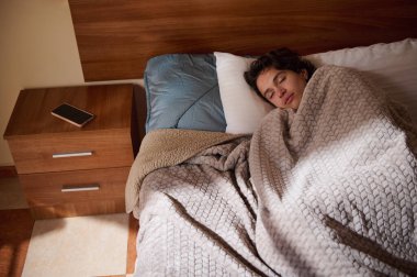 Huzurlu bir yatak odası sahnesinde genç bir çocuk yastıkla yumuşak bir battaniyenin altında uyurken telefon ahşap bir komodinin üzerinde dinleniyor, dinleniyor, rahatlıyor ve sessiz anlar yaşanıyor..