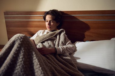 Genç bir kadın kalın dokulu bir battaniyenin altında uyuyor, okuyor ya da dinleniyor. Ilık ışıklandırma, ahşap yatak başlığı ve sakin, rahat bir ortam özel, samimi bir yer yaratıyor..